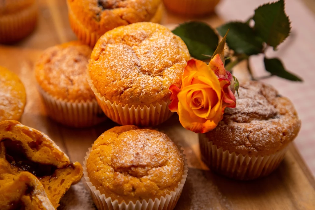 Kilka złocistych muffinek na drewnianej desce, oprószonych cukrem pudrem, ozdobionych pomarańczową różą, jedna babeczka rozkrojona pokazuje nadzienie.