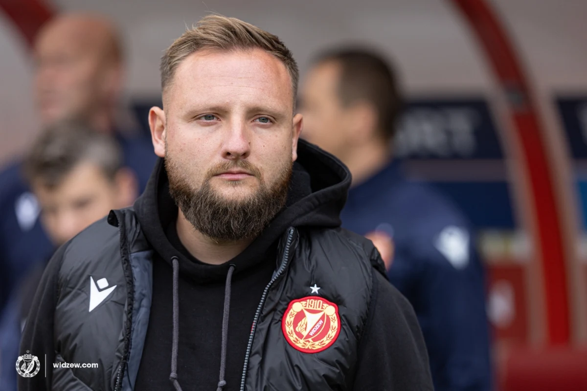 Mężczyzna z brodą w czarnej kurtce sportowej i bluzie z kapturem, z emblematem klubu piłkarskiego na piersi, stoi na tle rozmytych postaci i elementów stadionu.