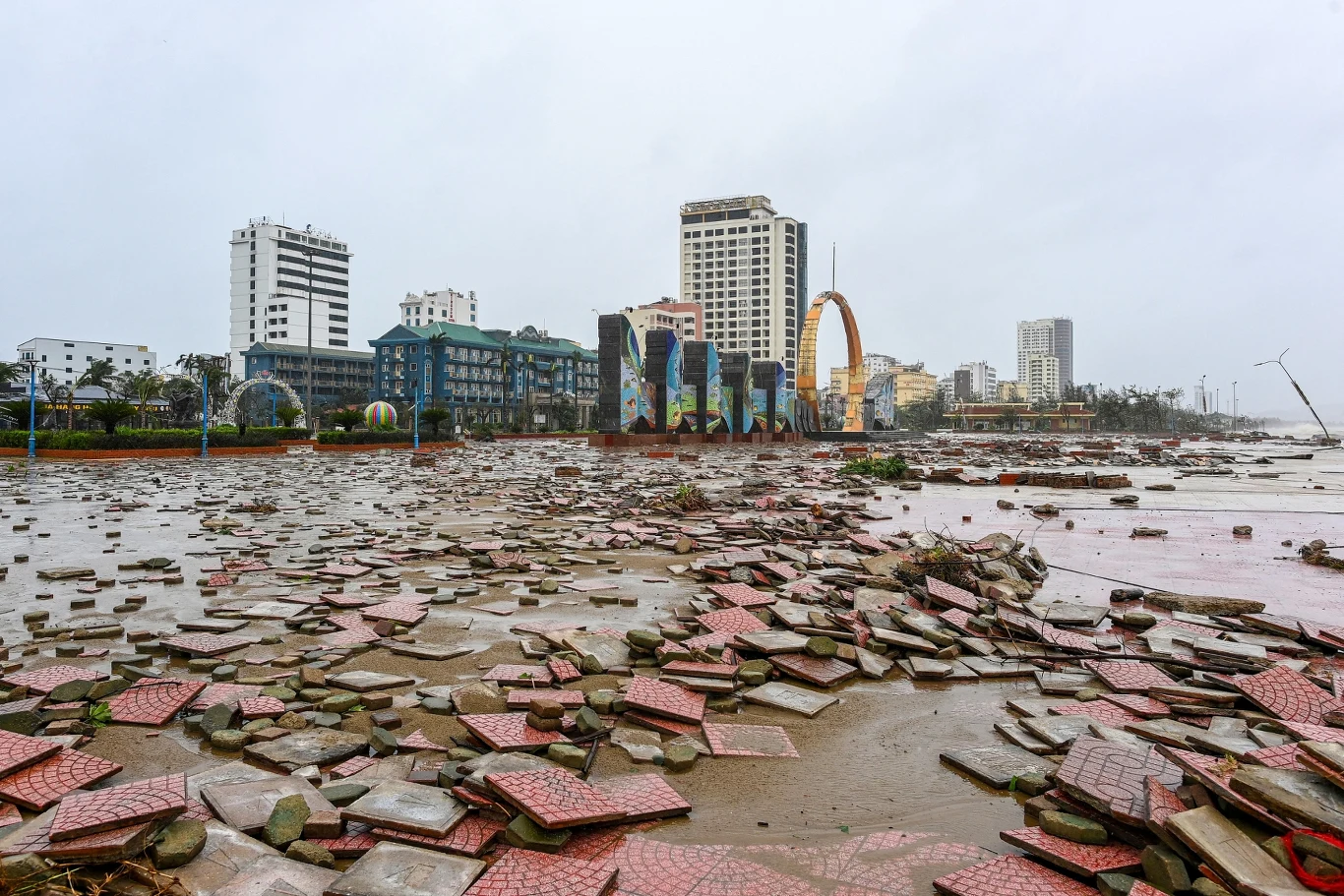 Plaża Cua Lo w prowincji Nghe An po przejściu przez Wietnam tajfunu Bualoi Zniszczony deptak nadmorski z rozrzuconymi płytami chodnikowymi, na drugim planie wysokie budynki i charakterystyczna brama, szare niebo i ślady ulewy sugerujące przejście gwałtownej burzy lub tajfunu.