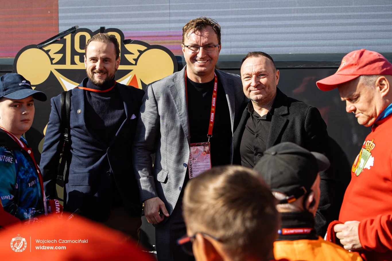 Przed meczem z Rakowem Robert Dobrzycki, głowny udziałowiec Widzewa spotkał się z kibicami Grupa mężczyzn pozuje do zdjęcia na tle dużego logo klubu sportowego, wokół widoczna jest także publiczność ubrana w ubrania nawiązujące do barw klubowych.