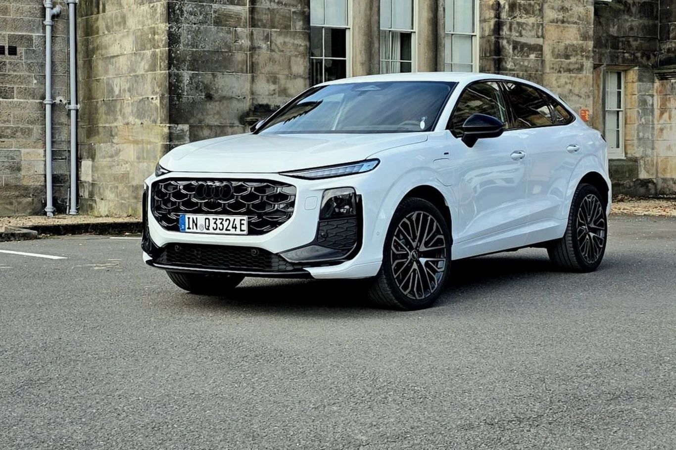 Audi Q3 posiada dwa warianty nadwozia: SUV oraz Sportback Biały nowoczesny samochód osobowy typu SUV stoi zaparkowany na tle zabytkowego, kamiennego budynku, wyróżnia się designerskimi reflektorami LED, dużymi felgami i masywnym grillem.