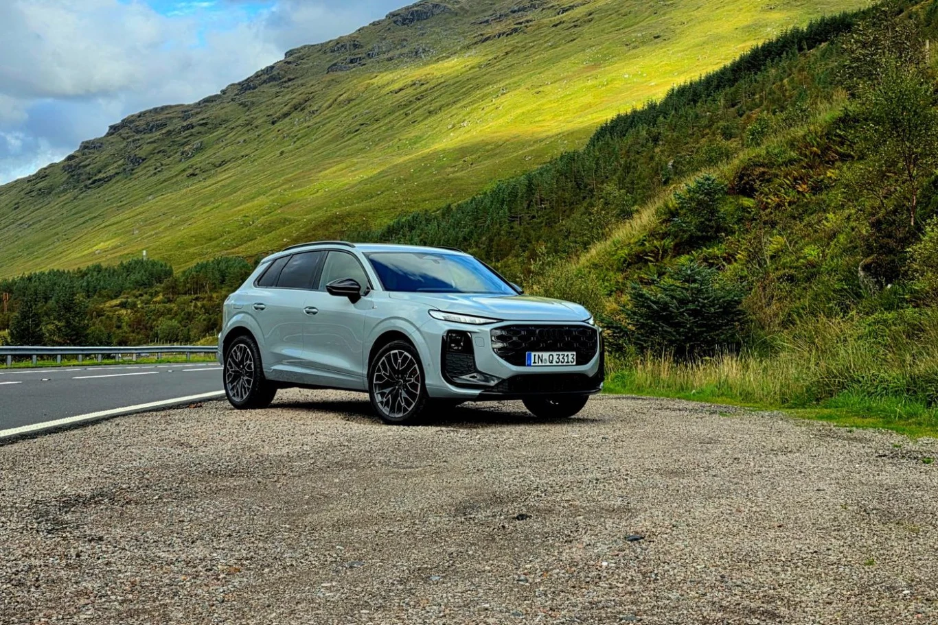 Audi Q3 SUV poznacie, po większym oknie za tylnymi drzwiami i prostszej linii dachu Srebrny SUV zaparkowany przy górskiej drodze, w tle zielone zbocza gór oraz niewielkie drzewa, podłoże pokryte żwirem, po lewej stronie fragment asfaltowej jezdni.