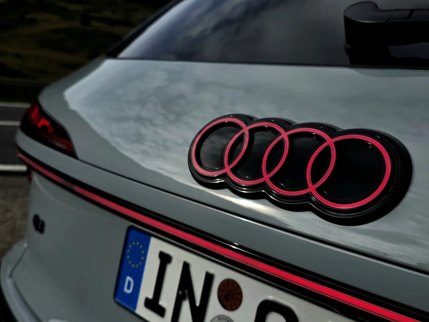 Podświetlony logotyp robi wrażenie po zmroku Logo marki Audi w kolorze czarnym z różowym konturem na tylnej części samochodu, widoczna część tylnej szyby oraz fragment niemieckiej tablicy rejestracyjnej.