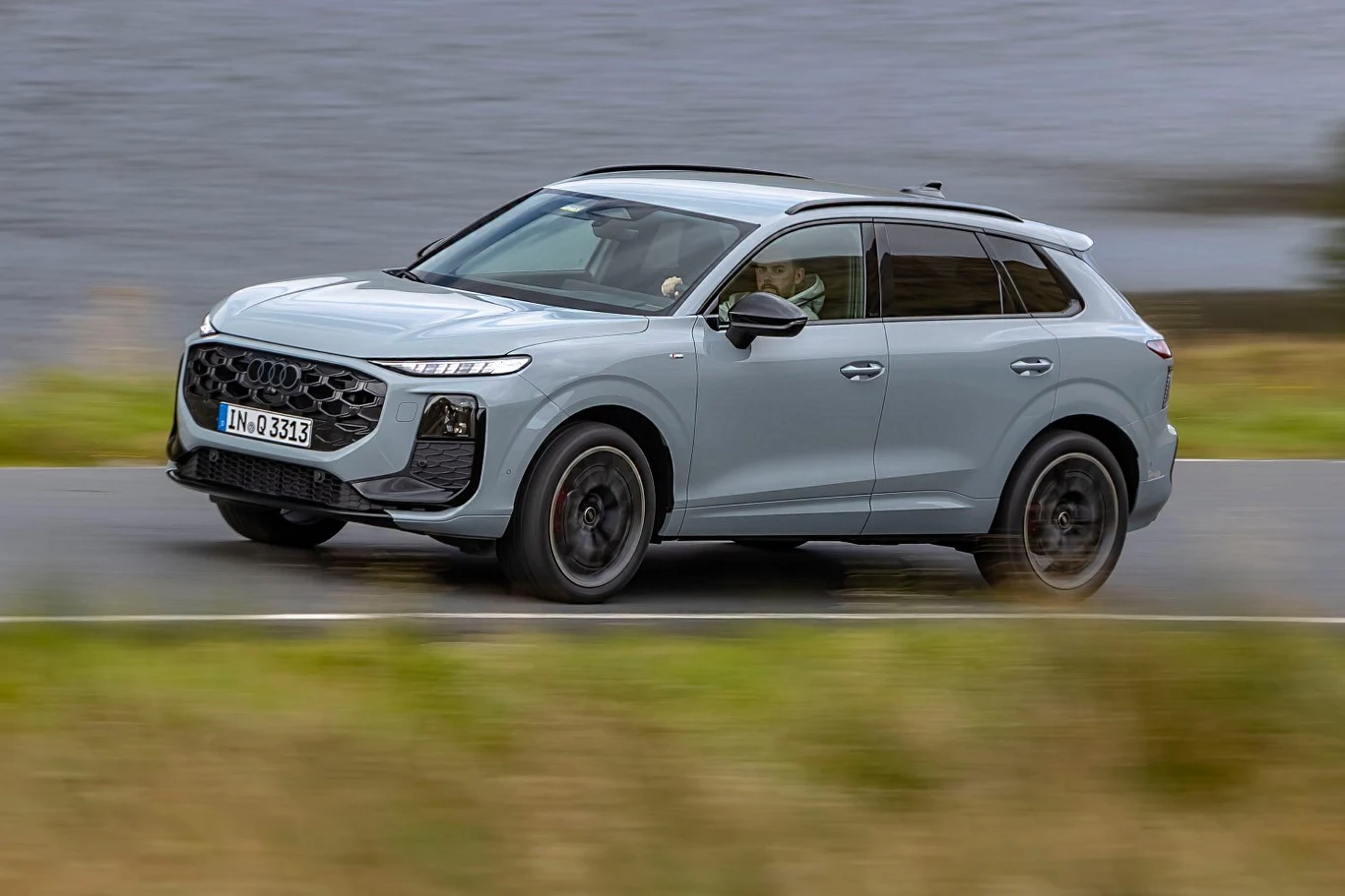Audi Q3 e-hybrid na prądzie przejedzie nawet 120 kilometrów Szary SUV jedzie z umiarkowaną prędkością po asfaltowej drodze w naturalnym krajobrazie, z tyłu widoczna rozmyta tafla wody i fragment zieleni.