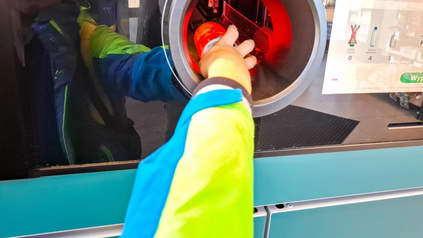System kaucyjny obejmuje puszki po napojach Ręka osoby umieszczającej pustą butelkę plastikową w automacie do odbioru opakowań zwrotnych, na ekranie widoczne są instrukcje użytkowania.