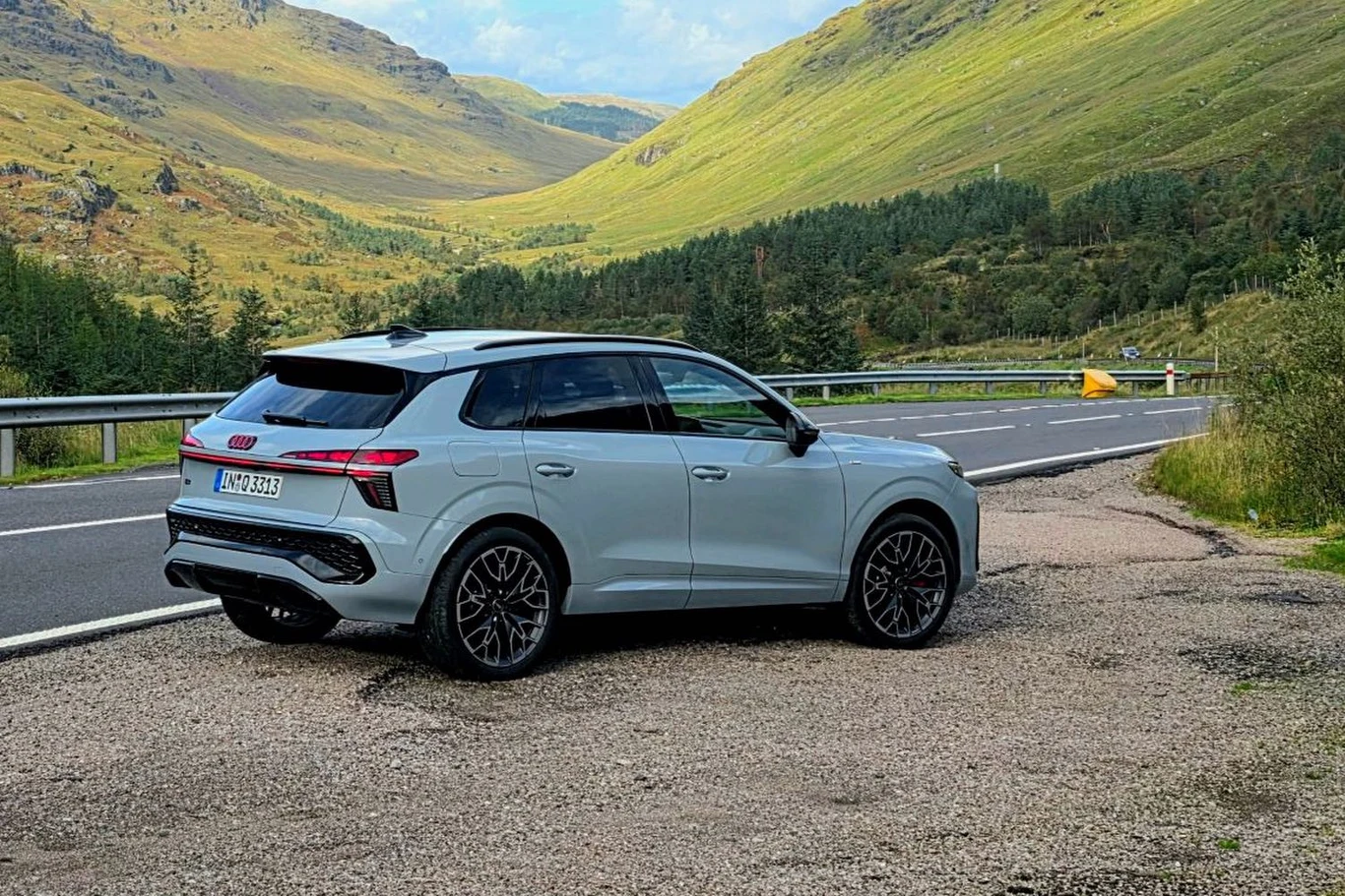 Najtaniej nowe Audi Q3 kupicie za 188 900 złotych Nowoczesny biały SUV marki Audi zaparkowany na poboczu asfaltowej drogi, otoczony zielonymi pagórkami i górami, w tle fragment lasu oraz górzysty krajobraz.