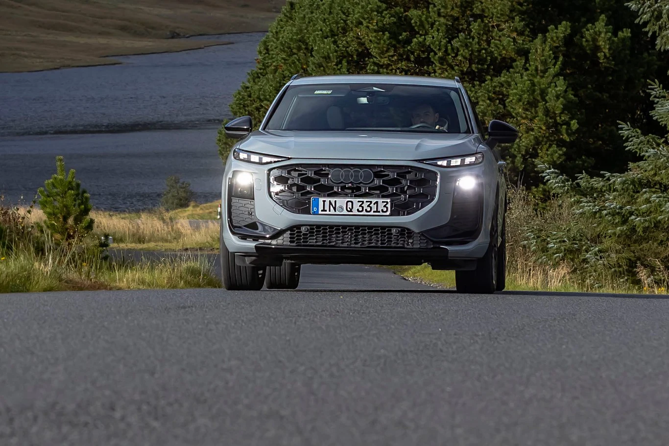 Nowe reflektory Audi Q3 składają się z 25 600 elementów Nowoczesny srebrny samochód osobowy marki Audi jedzie asfaltową drogą otoczoną zielenią oraz lasem, w tle widoczne jezioro i pagórkowaty krajobraz.