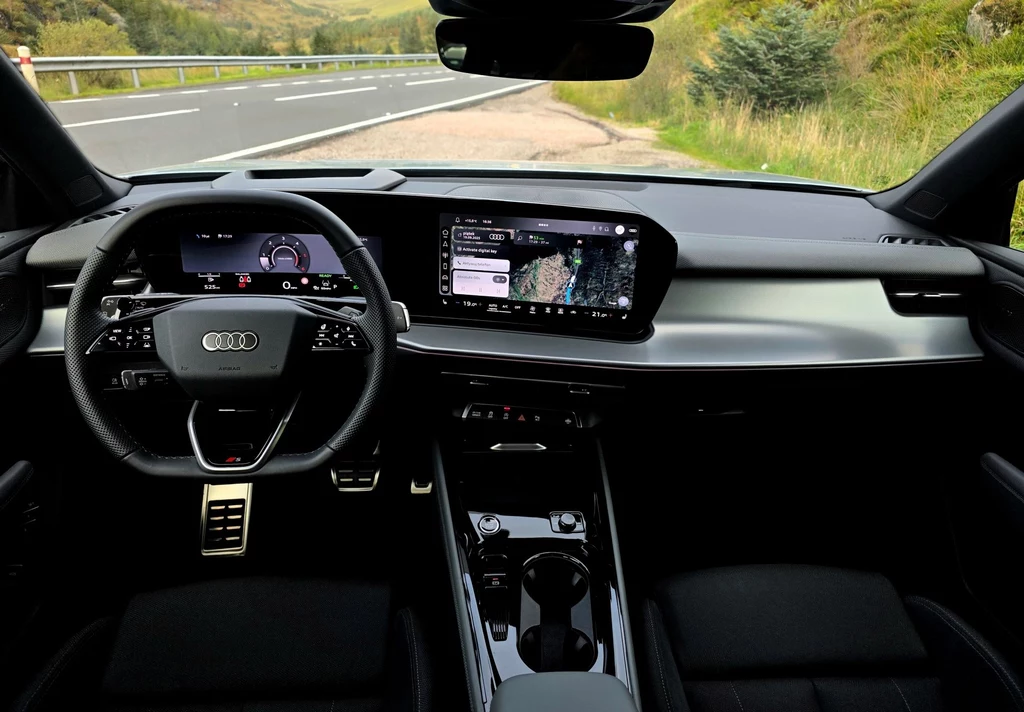 Wnętrze nowoczesnego samochodu marki Audi, z widocznym cyfrowym kokpitem, kierownicą wielofunkcyjną, panelem multimedialnym oraz wygodnymi siedzeniami, na tle otoczenia drogi oraz naturalnego krajobrazu za szybą.