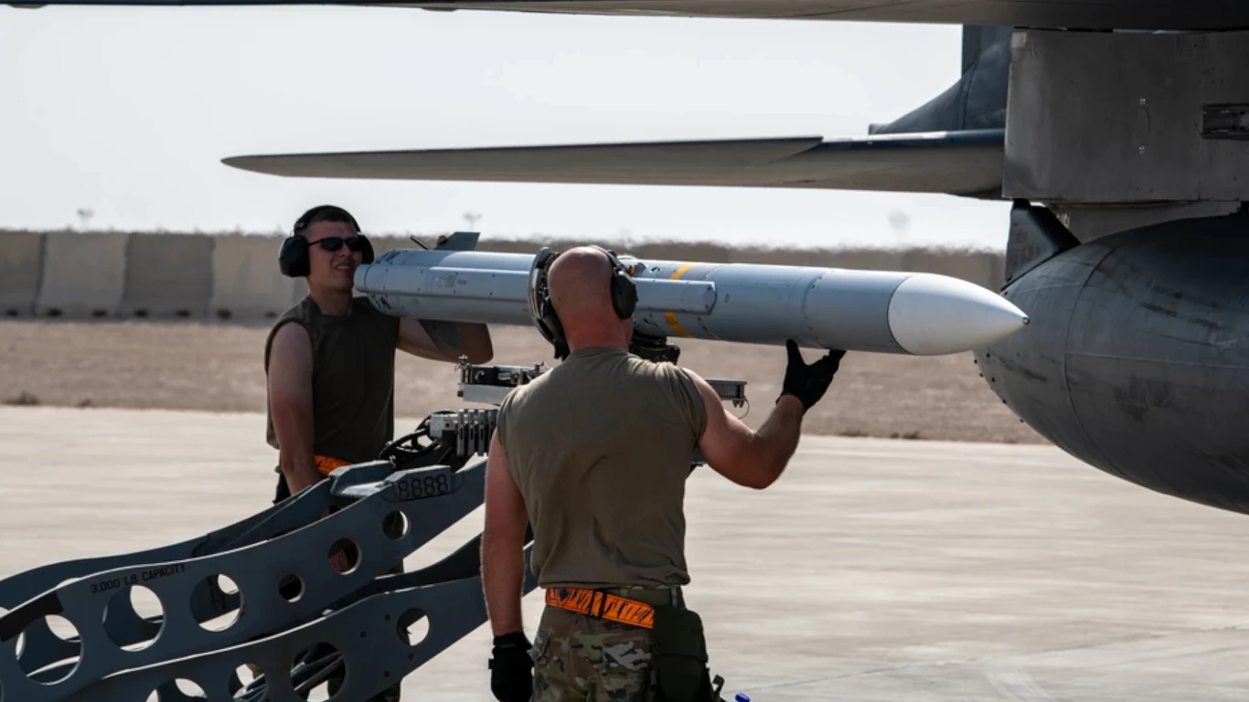 Amerykańcy żołnierze montują pocisk AIM-120D-3 pod myśliwiec F-15E Strike Eagle Żołnierze w mundurach i ochronnych słuchawkach umieszczają pocisk rakietowy pod skrzydłem samolotu wojskowego na betonowym pasie lotniska.