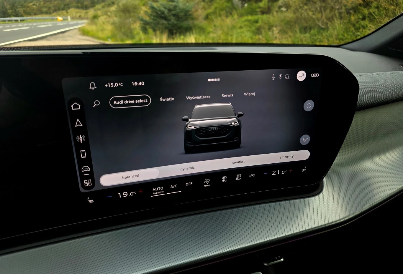 W Audi Q3, panel do obsługi klimatyzacji, to stała część centralnego ekranu Nowoczesny ekran multimedialny w samochodzie wyświetlający interfejs konfiguracji pojazdu, w centrum widoczny wizerunek samochodu marki Audi i opcje nawigacji systemowej, wokół panele sterowania klimatyzacją i innymi funkcjami auta.