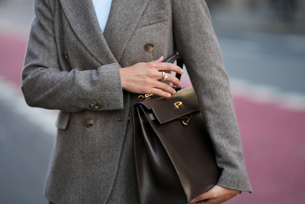 Elegancka osoba w szarej, dwurzędowej marynarce trzymająca stylową, brązową torebkę skórzaną z metalowymi zapięciami i pierścionkami na palcach.