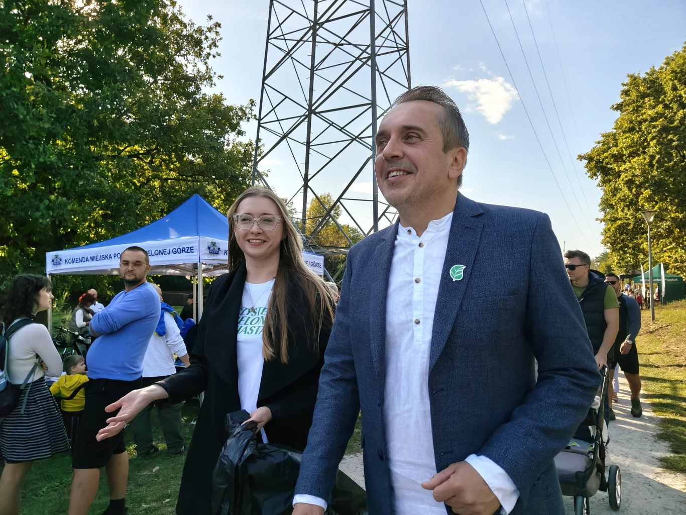 Uśmiechnięty mężczyzna w marynarce i kobieta w okularach idą obok siebie na tle grupy ludzi oraz biało-niebieskiego namiotu. W tle widoczna wieża energetyczna, drzewa i stoiska eventowe.