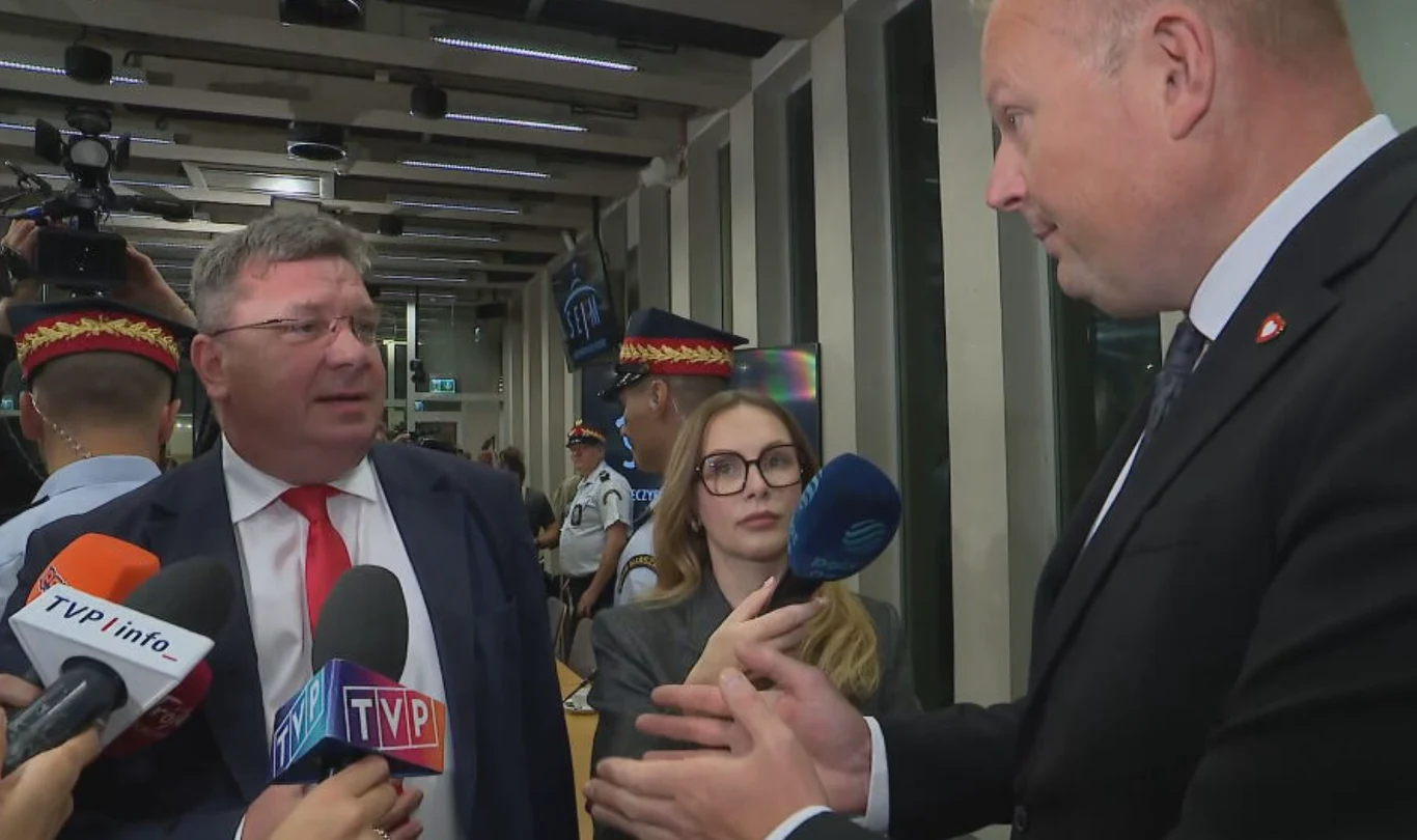 Spięcie polityków przed przesłuchaniem Zbigniewa Ziobry Dwóch mężczyzn ubranych w garnitury rozmawia, otoczonych przez dziennikarzy trzymających mikrofony różnych stacji telewizyjnych, na drugim planie widoczni są funkcjonariusze w odświętnych mundurach.