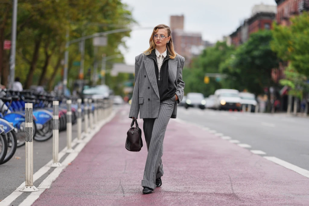 Kobieta w modnym, szarym garniturze w paski idzie środkiem miejskiej ulicy, trzymając w ręce ciemną torbę, w tle widoczne są drzewa, zaparkowane rowery i miejskie budynki.