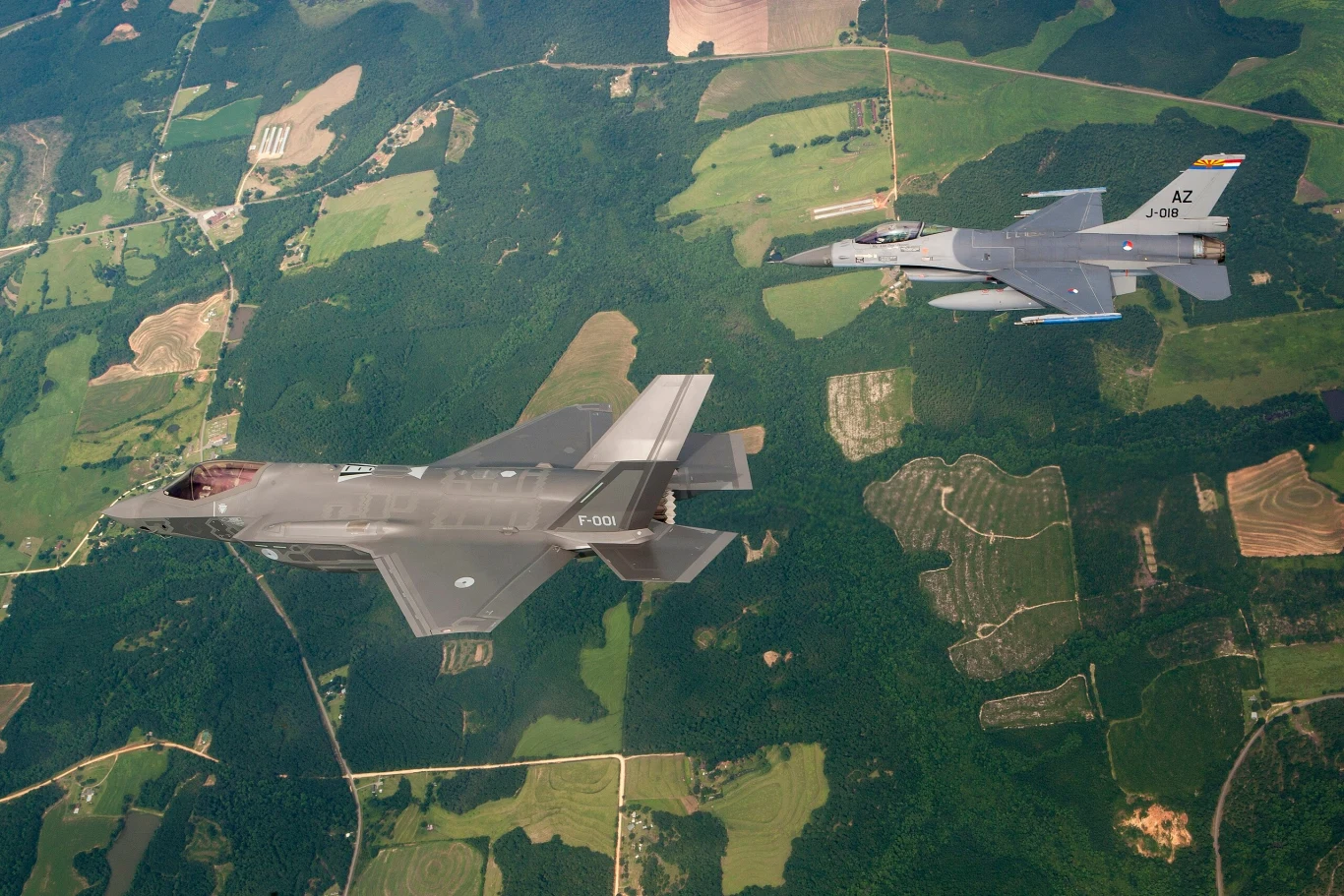 W polskich siłach powietrznych pociski AIM-120 w wersjach C mają być używane przede wszystkim przez F-16. F-35 ma także przenosić je w wersji D, jako broń specjalna Dwa myśliwce wojskowe lecące nad zielono-brązowym krajobrazem z polami uprawnymi i lasami, jedno z nich to F-35, drugie F-16, widok z góry, oba samoloty znajdują się w locie szybowym.
