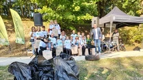 Grupa ludzi pozuje z torbami i nagrodami pod namiotem oraz flagami organizatora, przed nimi leżą wypełnione worki na śmieci i stara opona jako dowód zakończonej akcji sprzątania terenów zielonych.