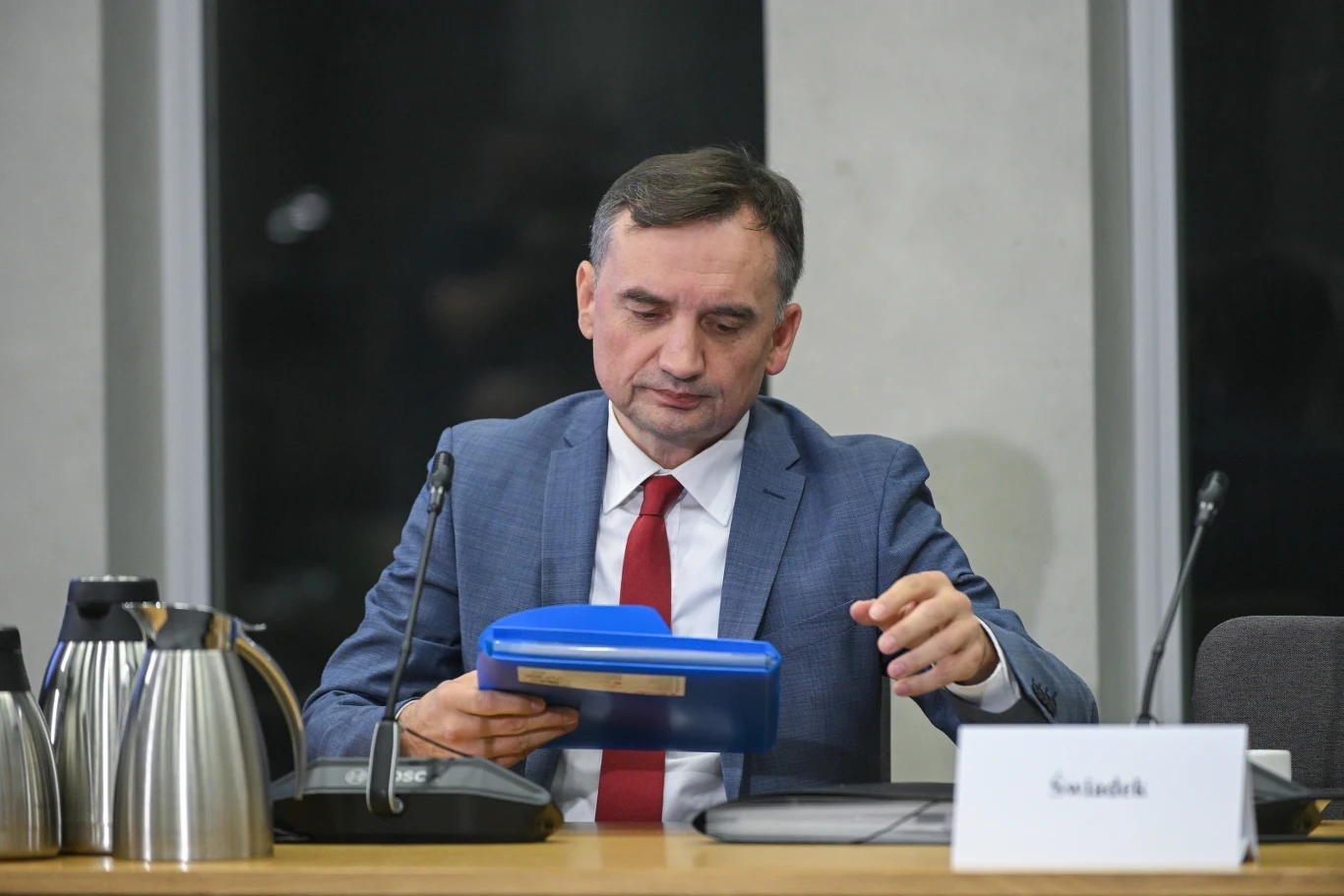Zbigniew Ziobro przed komisją śledczą ds. Pegasusa Mężczyzna w garniturze i czerwonym krawacie siedzi przy stole konferencyjnym, trzyma w rękach niebieski segregator, na stole znajdują się dzbanki i mikrofony, a przed nim stoi tabliczka z napisem 'Świadek'.