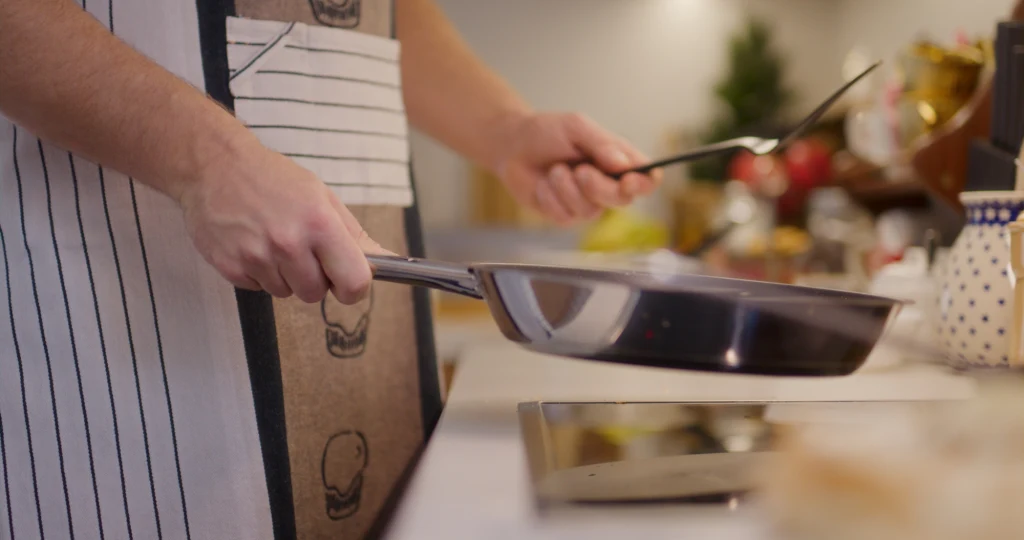 Jak zrobić tosty bez chleba? osoba w fartuchu kuchennym trzyma patelnię nad płytą grzewczą i używa łopatki, kuchnia jest dobrze oświetlona, w tle widać różne naczynia i składniki