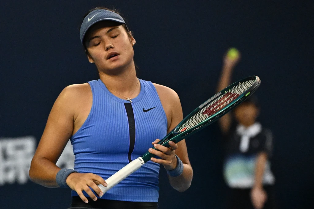 Emma Raducanu Tenisistka w niebieskim stroju sportowym stojąca z zamkniętymi oczami i opartą rakietą tenisową o ramię, w tle rozmyty sędzia lub chłopiec podający piłki.