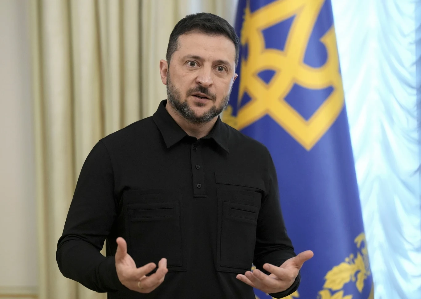 Prezydent Ukrainy Wołodymyr Zełenski Mężczyzna w czarnej koszuli gestykuluje stojąc przed dużą niebieską flagą z żółtym herbem, w tle jasne zasłony.
