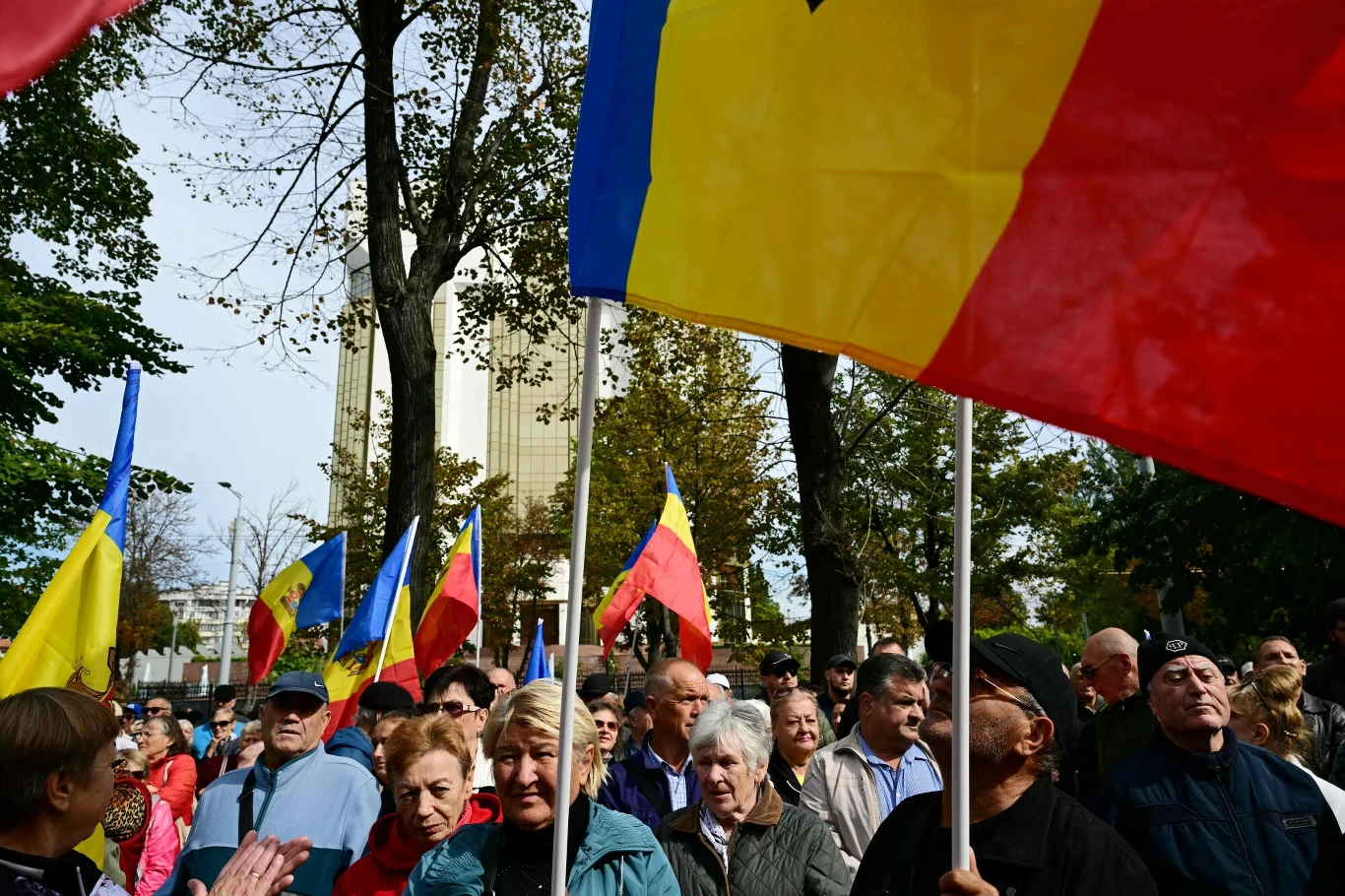 Wybory w Mołdawii wygrały najpewniej siły proeuropejskie Tłum ludzi na zewnątrz, wiele osób trzymających żółto-czerwono-niebieskie flagi, drzewa i budynek w tle, atmosfera pokojowego zgromadzenia.