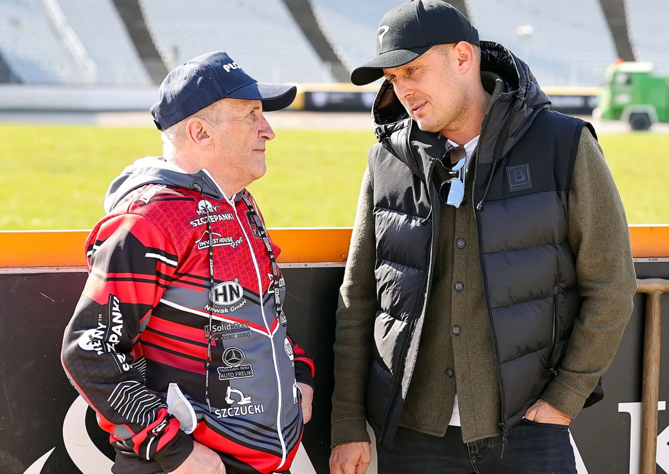 Dwóch mężczyzn rozmawia ze sobą na tle stadionu sportowego, jeden z nich ubrany w sportową kurtkę i czapkę, drugi w kamizelkę i czapkę z daszkiem, w tle widoczne puste trybuny oraz fragment trawy.