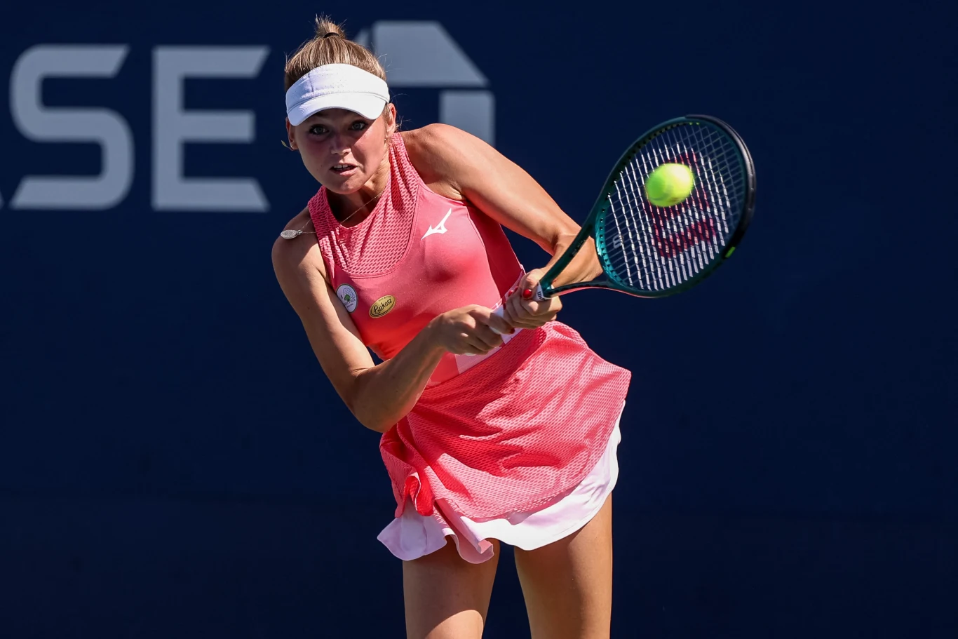 Tenisistka wykonuje zamach rakietą podczas meczu na otwartym korcie, ubrana w różową sukienkę sportową i białą czapkę z daszkiem, piłka tenisowa znajduje się w powietrzu.