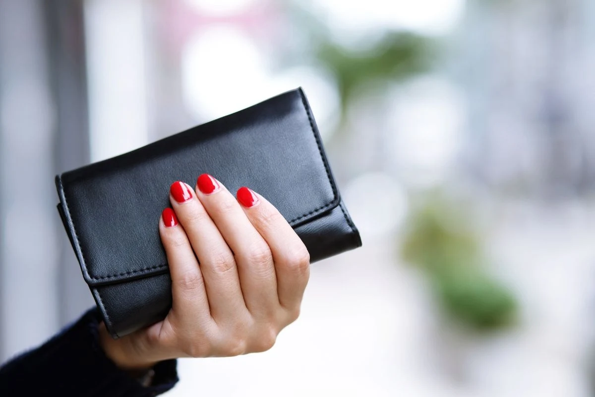 Dłoń z pomalowanymi na czerwono paznokciami trzyma czarny portfel na tle rozmytego tła.