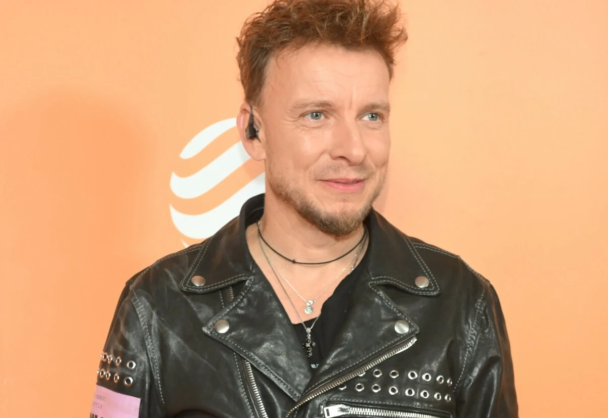 Piotr Kupicha utrzymuje dobry kontakt z synami Mężczyzna w skórzanej kurtce z ćwiekami, z krótkimi, zaczesanymi do góry włosami, lekko uśmiechający się na tle pomarańczowej ścianki z białym graficznym wzorem. Ma na szyi cienki naszyjnik i słuchawkę w uchu.