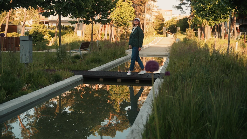 Kobieta idzie przez niewielki, drewniany mostek nad płytkim kanałem wodnym w otoczeniu zieleni oraz nowoczesnej zabudowy, otoczona drzewami i trawami przy ścieżce spacerowej.
