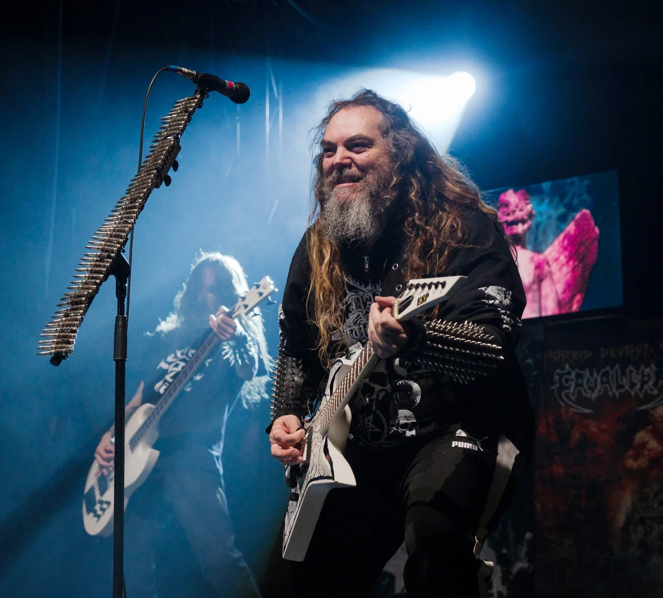 Max Cavalera stoi na czele grupy Soulfly, po lewej w tle Igor Amadeus Cavalera Muzyk z długimi włosami i brodą gra na białej gitarze elektrycznej na scenie, ubrany w czarną kurtkę z ćwiekami, obok drugi gitarzysta w tle, światło reflektora podkreśla dynamikę koncertu, na scenie widoczny ozdobiony mikrofon z nabojami.