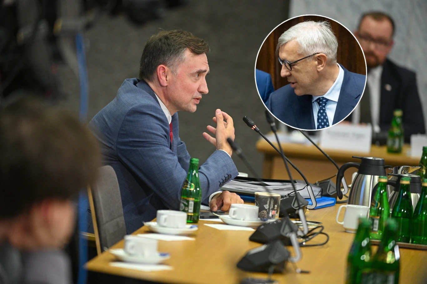 Zbigniew Ziobro zeznaje na komisji śledczej ds. Pegasusa, a Roman Giertych składa zawiadomienie Mężczyzna w garniturze gestykuluje podczas dyskusji przy stole konferencyjnym, wokół ustawione są butelki z wodą, filiżanki oraz notatki. W okrągłym wycinku widoczny inny mężczyzna w rozmowie, obaj biorą udział w formalnym spotkaniu.