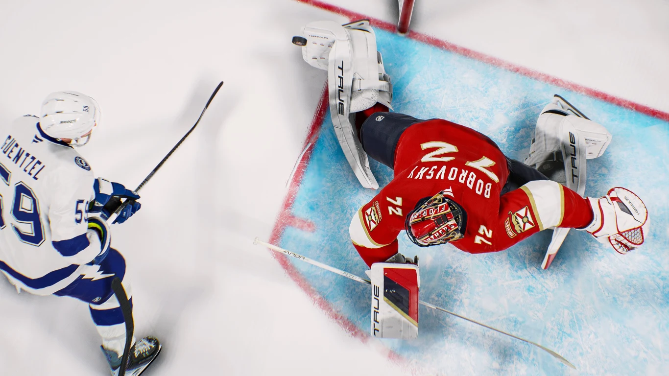 NHL 2K26 Bramkarz w czerwonym stroju wykonuje dynamiczną obronę na lodowisku, podczas gdy zawodnik drużyny przeciwnej w białej koszulce i niebieskich spodenkach zmierza w stronę bramki, trzymając kij hokejowy.