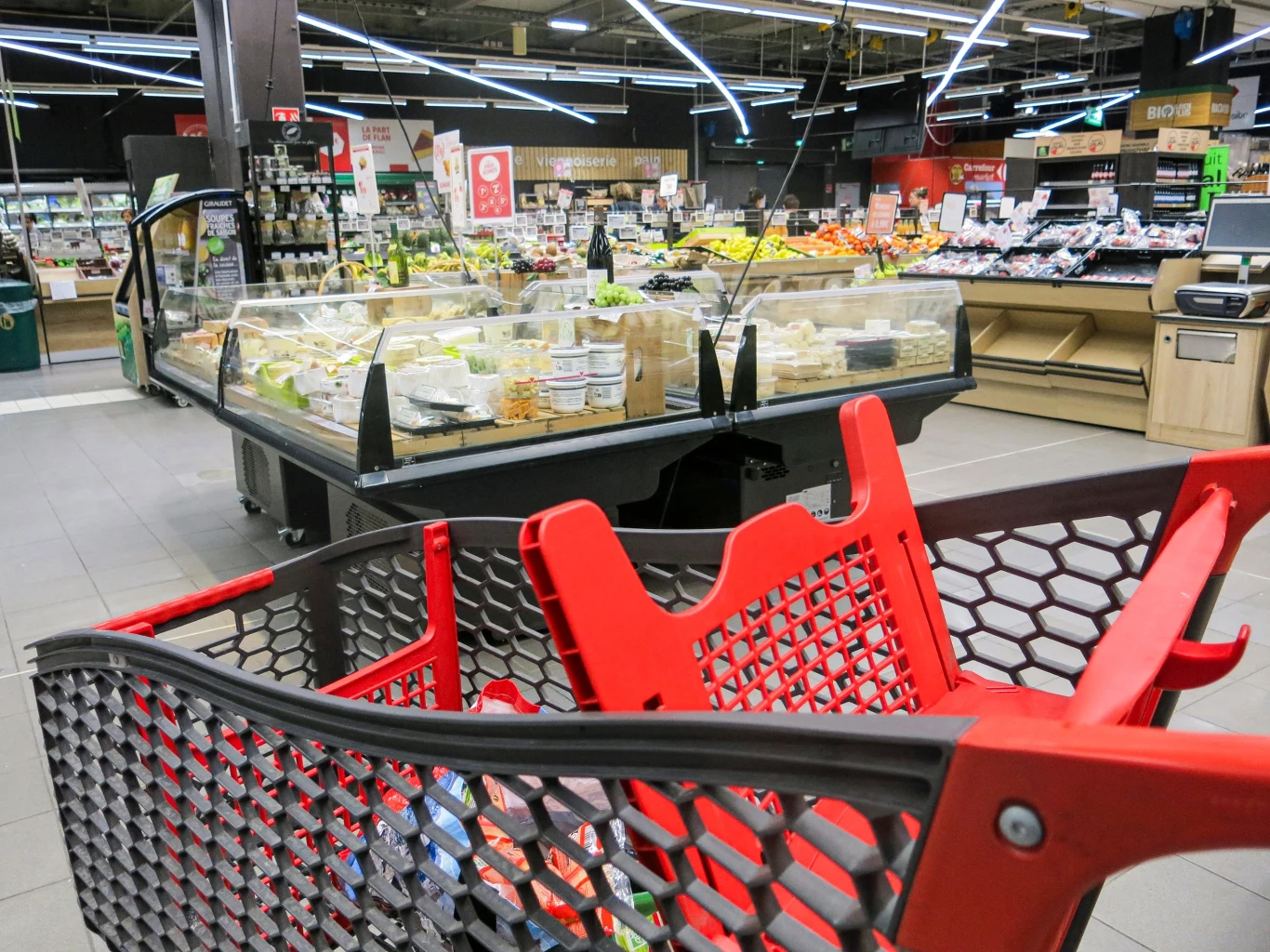 Francuskie media: Carrefour sonduje możliwość sprzedania aktywów w Polsce Wnętrze nowoczesnego supermarketu z czerwonym wózkiem zakupowym na pierwszym planie, w tle dobrze zaopatrzona lada z nabiałem oraz warzywami i owocami, jasne oświetlenie podkreśla estetykę i porządek przestrzeni handlowej.