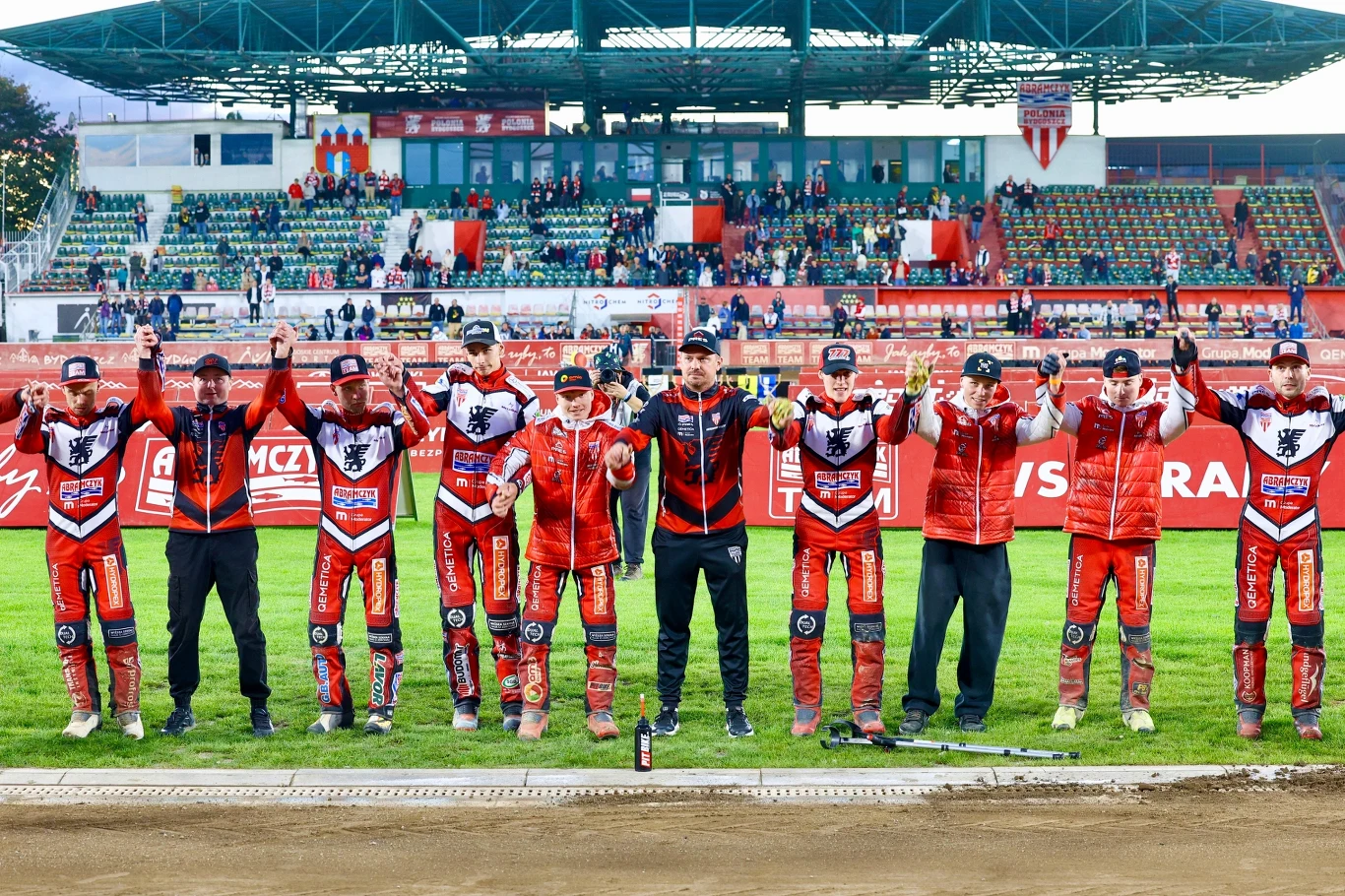 Drużyna żużlowa ubrana w czerwono-czarne stroje stoi ramię w ramię na murawie stadionu, uniesione ręce tworzą gest zwycięstwa, w tle widoczna trybuna z kibicami.