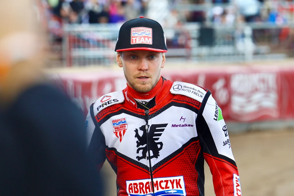 Zawodnik żużlowy w stroju sportowym z logotypami sponsorów, skupiony na twarzy, stoi na arenie żużlowej, w tle rozmyty tłum kibiców oraz ogrodzenie stadionu.