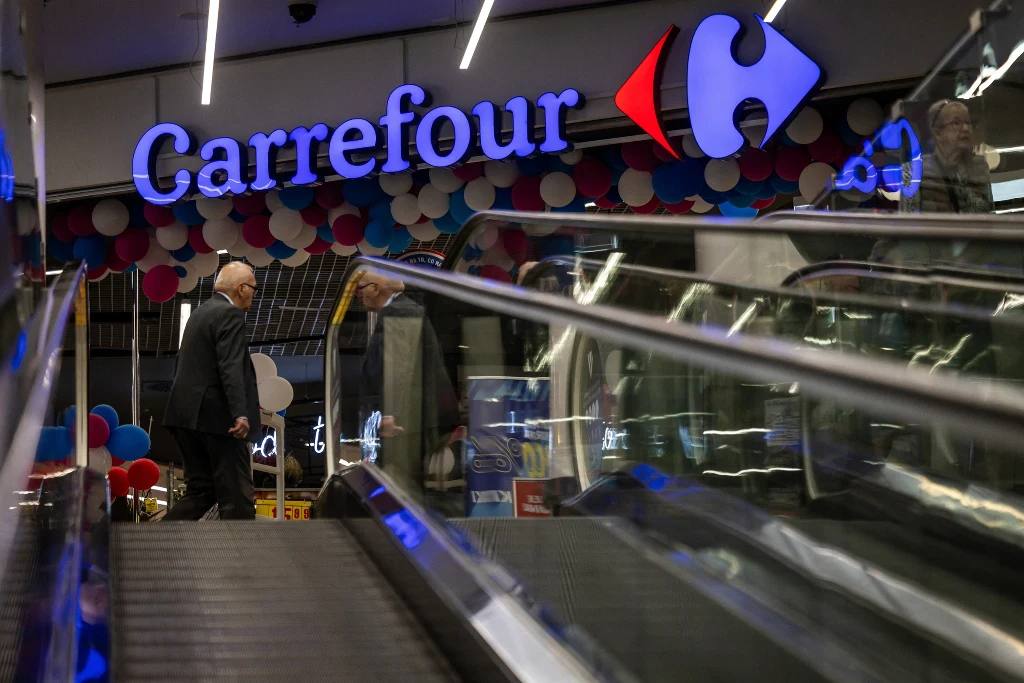 Widoczne wejście do supermarketu Carrefour, w tle oświetlone logo marki oraz dekoracje z balonów, osoby korzystają z ruchomych schodów.
