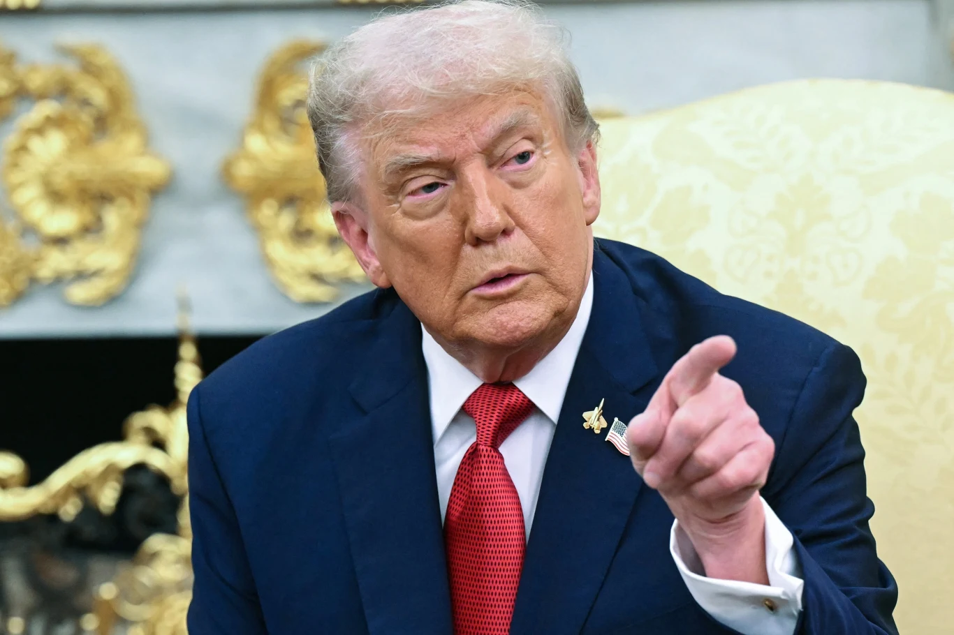 YouTube zapłaci Trumpowi gigantyczną ugodę Mężczyzna w garniturze z czerwoną krawatą siedzi na kremowym fotelu w ozdobnym wnętrzu, wskazując palcem w bok i patrzy w tym samym kierunku, z poważnym wyrazem twarzy.