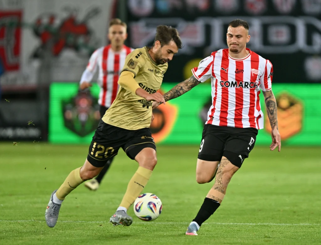 Dwóch piłkarzy rywalizuje o piłkę na murawie podczas meczu, jeden z nich w złotej koszulce wykonuje dynamiczny zwód, podczas gdy drugi w biało-czerwonych pasach próbuje odebrać piłkę.