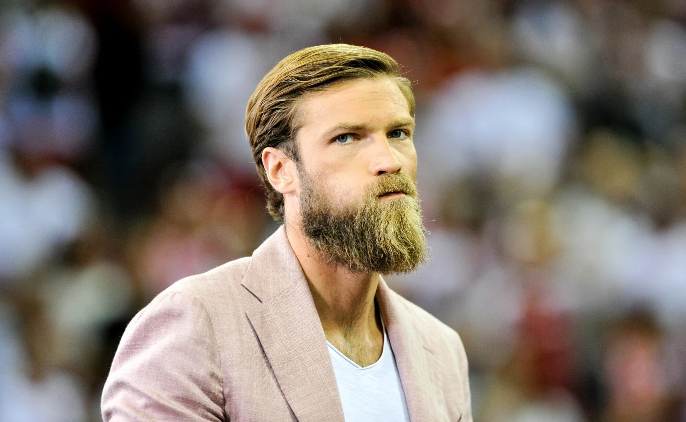 Brodaty mężczyzna w jasnej marynarce i białej koszulce na tle rozmytego tłumu w jasnych ubraniach, poważny wyraz twarzy.