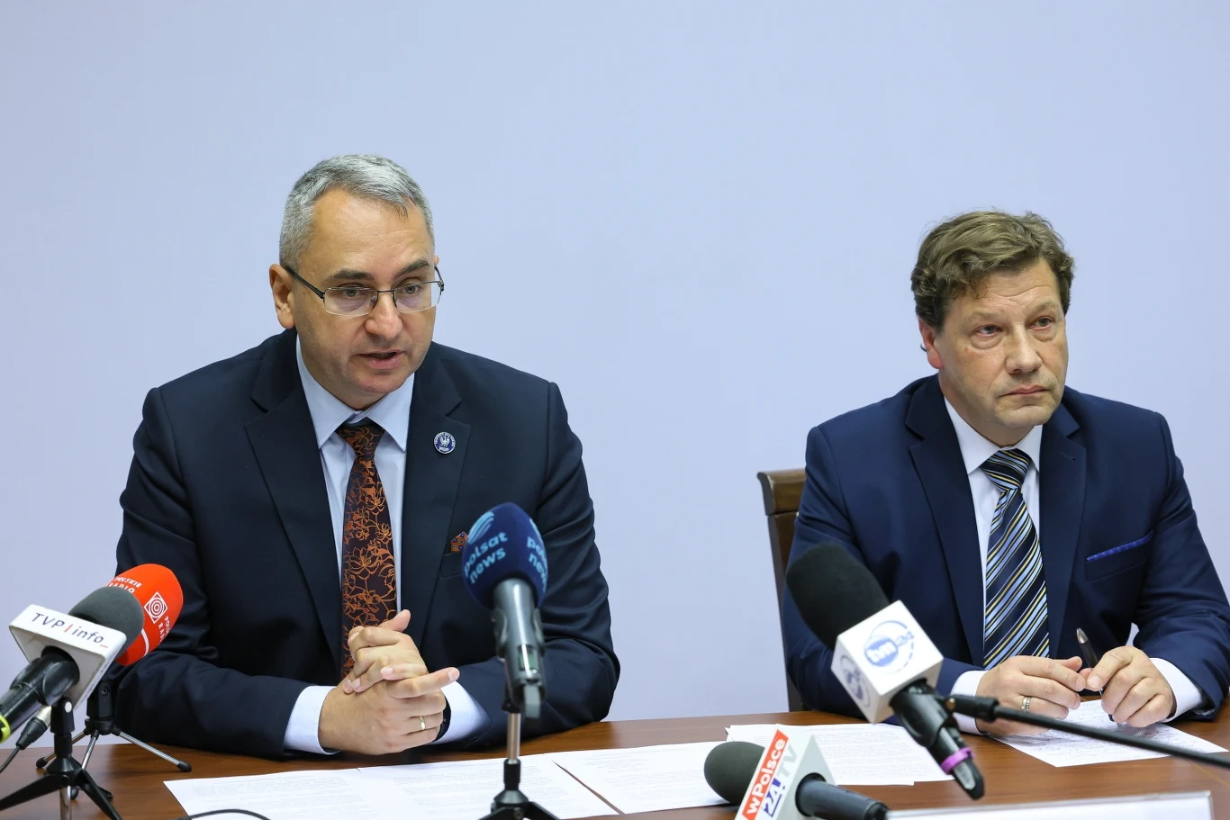 Sędzia Michał Lasota oraz sędzia Piotr Schab Dwóch mężczyzn w garniturach siedzi przy stole konferencyjnym z mikrofonami i dokumentami, jeden z nich mówi, drugi sporządza notatki, tło jasne i neutralne.