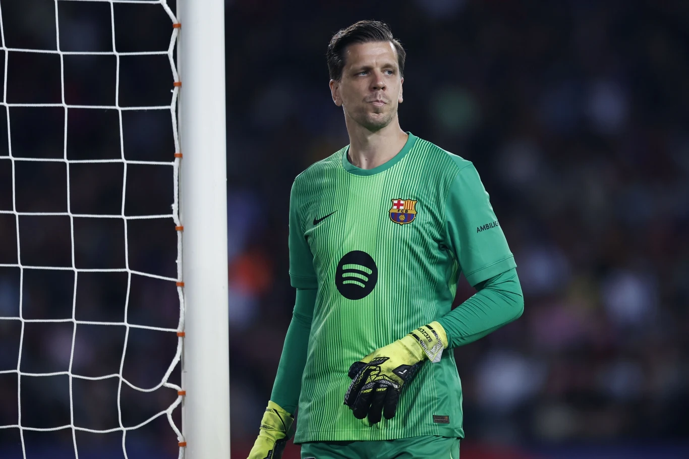 Bramkarz w zielonym stroju z logo klubu FC Barcelona oraz sponsora Spotify stoi obok słupka bramki na tle rozmytej publiczności stadionu, trzymając rękawice bramkarskie.