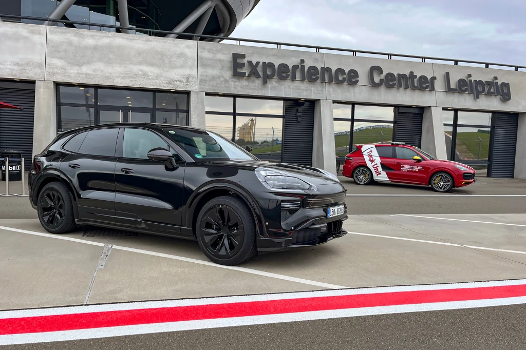 Dwa samochody marki Porsche zaparkowane przed budynkiem Experience Center Leipzig, z których jeden jest czarny SUV, a drugi czerwony sportowy samochód, widoczny asfalt oraz napisy na elewacji.