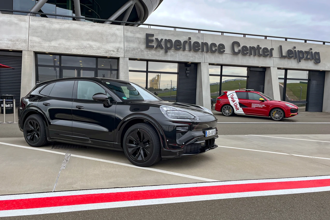 Dwa samochody marki Porsche zaparkowane przed budynkiem Experience Center Leipzig, z których jeden jest czarny SUV, a drugi czerwony sportowy samochód, widoczny asfalt oraz napisy na elewacji.