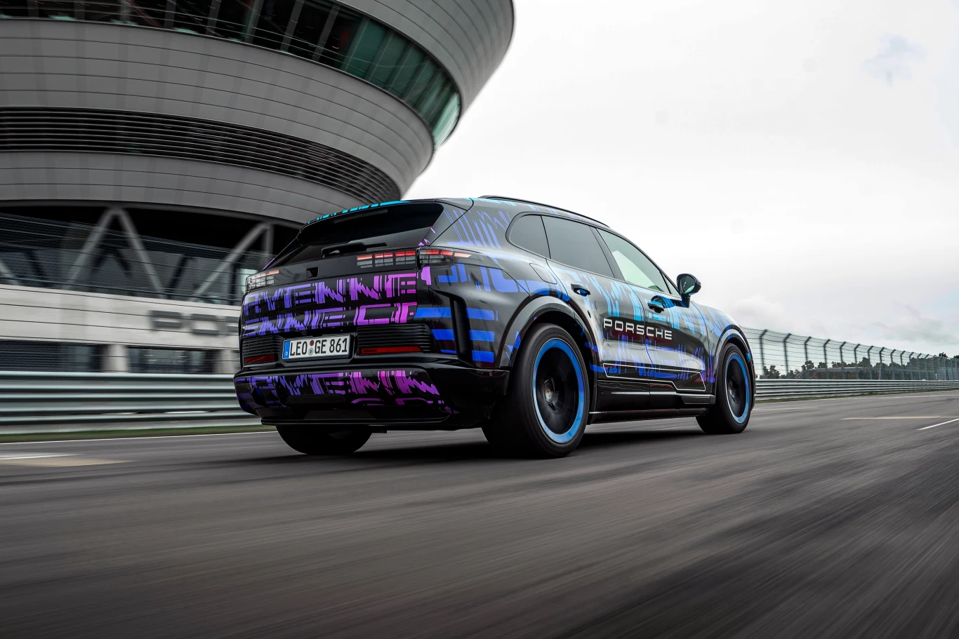 Nowoczesny SUV marki Porsche w ruchu na torze testowym, dynamiczna sylwetka pojazdu z futurystycznym wzorem karoserii na tle toru i nowoczesnego budynku.
