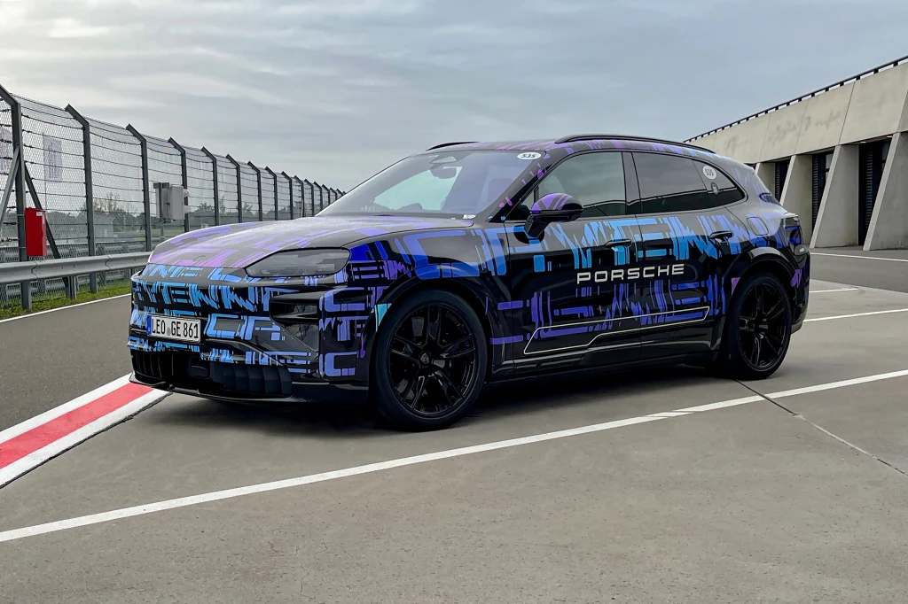 Koncepcyjny samochód Porsche w kamuflażu testowym, stojący na torze wyścigowym obok garaży, otoczony barierkami, charakterystyczny nowoczesny wygląd SUV-a z ciemnymi felgami