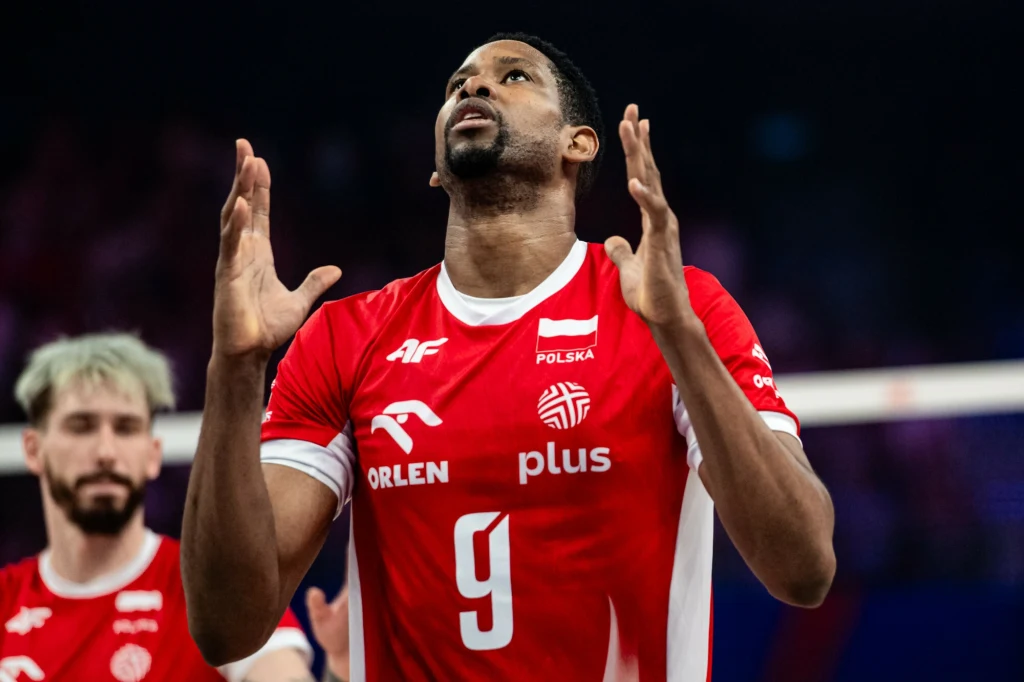 Siatkarz w czerwonym stroju reprezentacji Polski z numerem 9 na koszulce unosi dłonie w górę, za jego plecami widoczny jest inny zawodnik drużyny na tle sportowej hali.