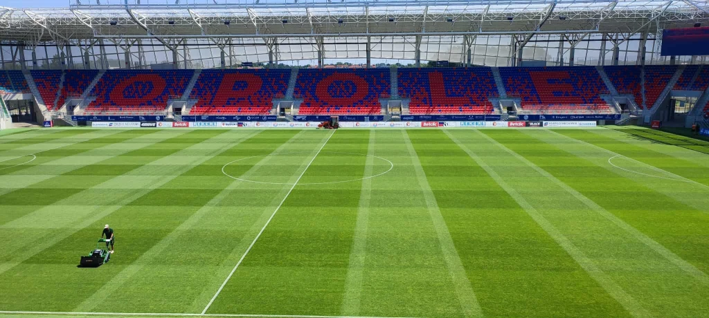 Trybuny stadionu piłkarskiego z napisem Opole, zielona murawa z wyraźnie zaznaczonymi pasami i osobą koszącą trawę.