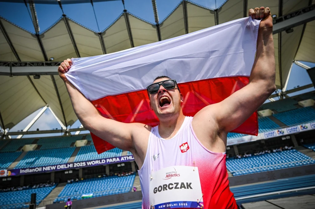Sportowiec w okularach przeciwsłonecznych trzyma wysoko nad głową biało-czerwoną flagę Polski na tle trybun stadionu, ubrany w oficjalny strój reprezentacyjny