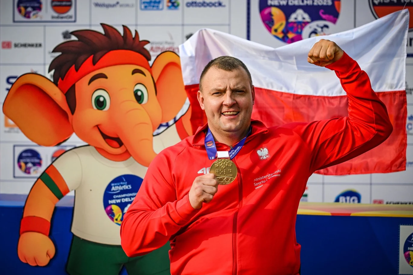 Bartosz Górczak już ze złotym medalem na szyi Sportowiec w czerwonym dresie z medalem na szyi, unosi zaciśniętą pięść w geście triumfu, w tle maskotka imprezy sportowej, polska flaga oraz tablica sponsorów.